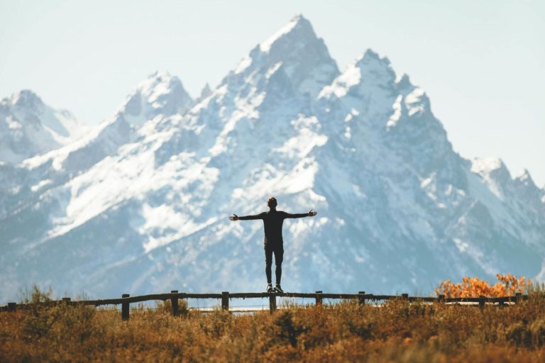 man in active wear with arms spread wide standing on a fence facing scenic snowy mountains